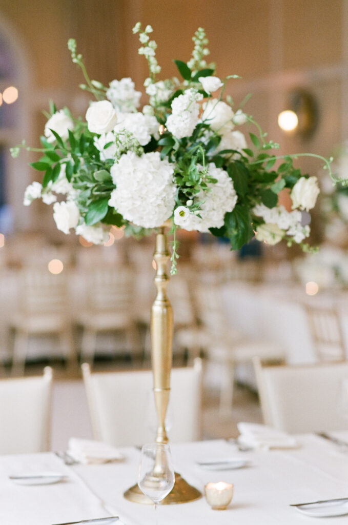 wedding-at-the-mount Elegant white floral centerpiece with hydrangeas and roses in a gold vase at a Wedding at The Mount, set in a classic reception space with ivory chairs and soft candlelight.