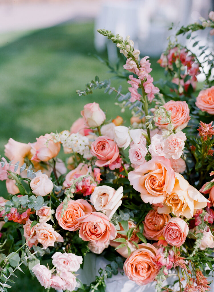 wedding-at-the-mount Close-up of a lush floral arrangement featuring peach, blush, and coral roses mixed with greenery and delicate pink snapdragons, set outdoors on a sunny day.