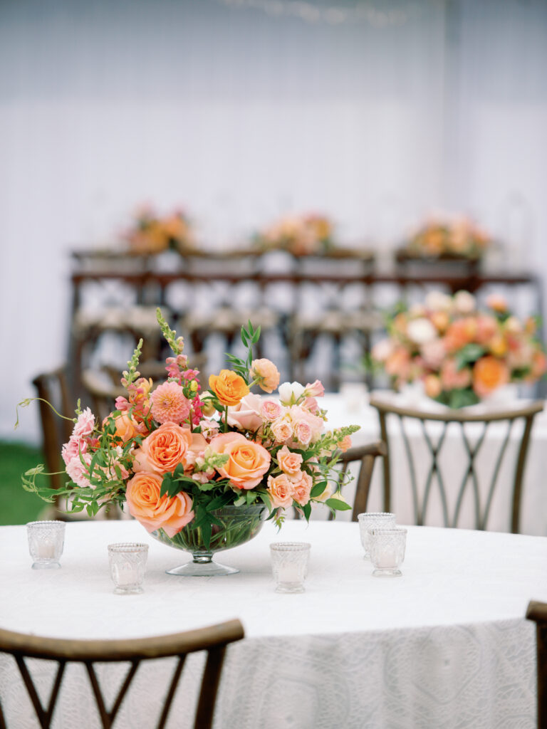 wedding-at-the-mount Lush centerpiece of orange and pink roses with greenery on a round table with white linen at a tented Wedding at The Mount.