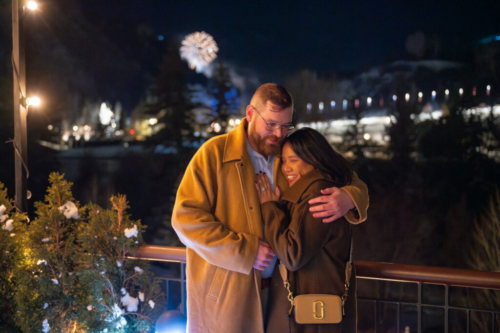 Engaged couple shares an intimate winter moment under fireworks on a snowy balcony at night.