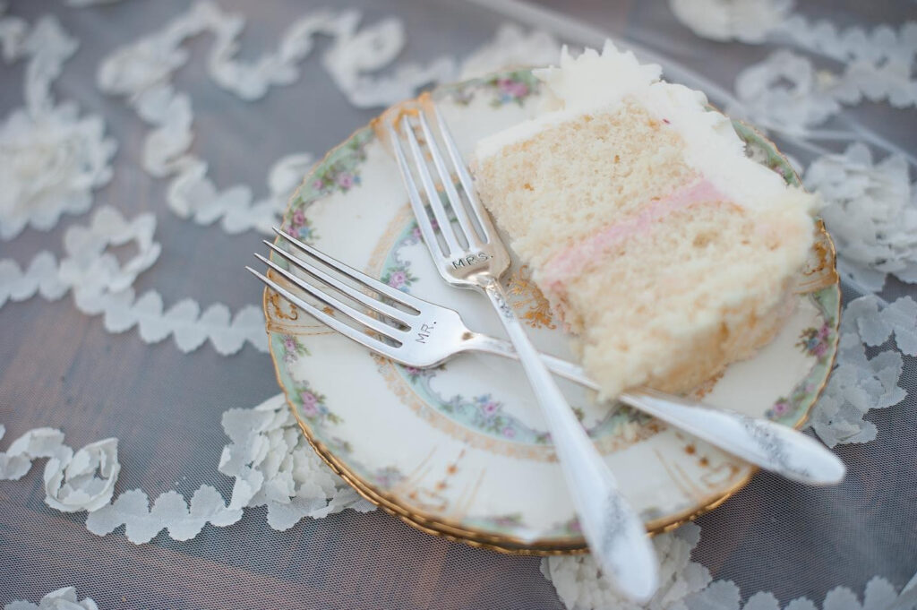 Slice of wedding cake on vintage china with “Mr.” and “Mrs.” forks, styled on delicate lace.