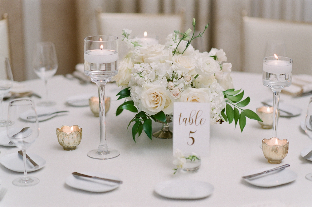 Elegant wedding table with white roses, floating candles, gold votives, and a "Table 5" card on a white tablecloth.

the newbury boston wedding