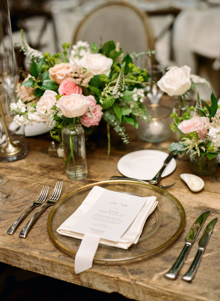 Rustic wedding table with floral centerpieces, clear charger, ivory napkin, printed menu, and candles in glass votives.

