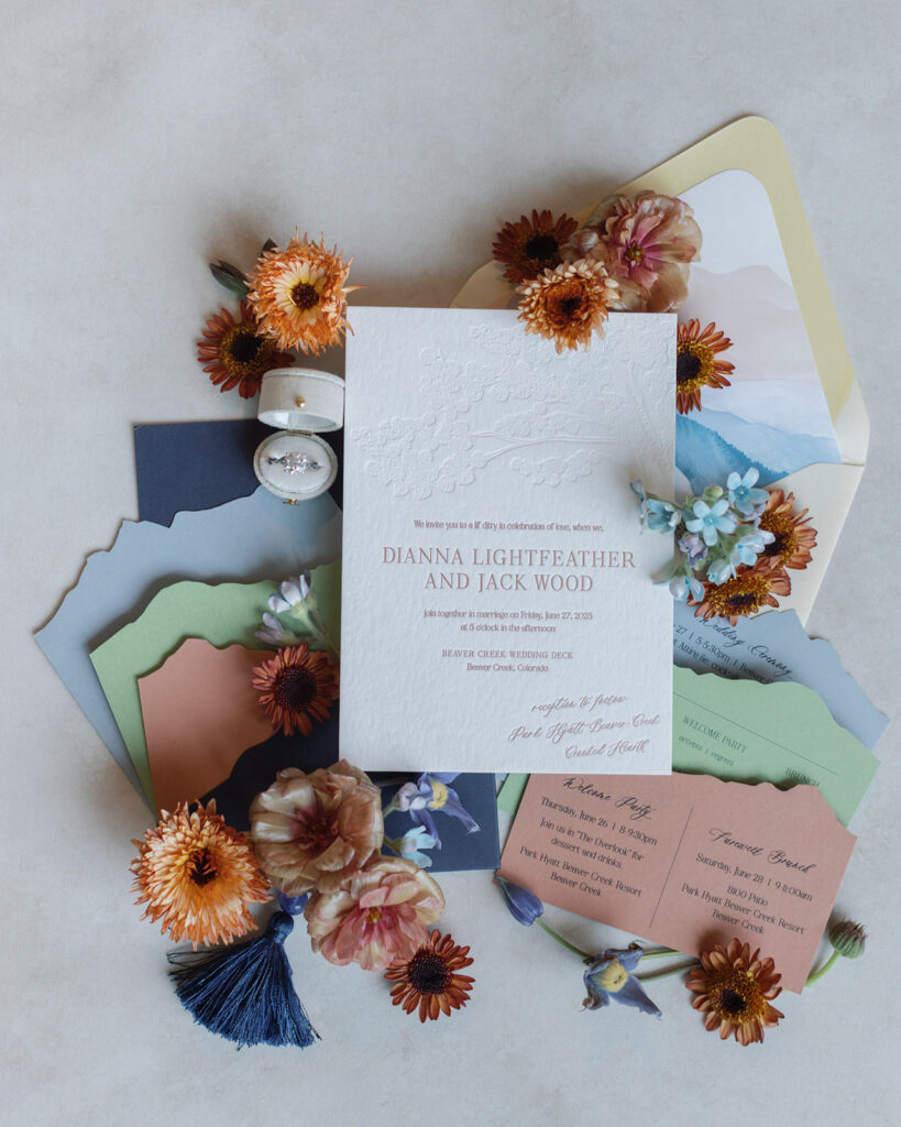 Elegant embossed invitation with colorful envelopes, orange and pink flowers, a diamond ring, and tassels arranged artfully on a neutral background.