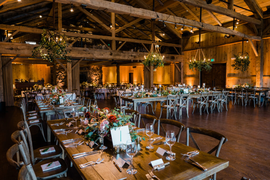 Wooden beams, long farm tables with colorful floral centerpieces, hanging greenery chandeliers, and warm golden lighting create a cozy wedding atmosphere.