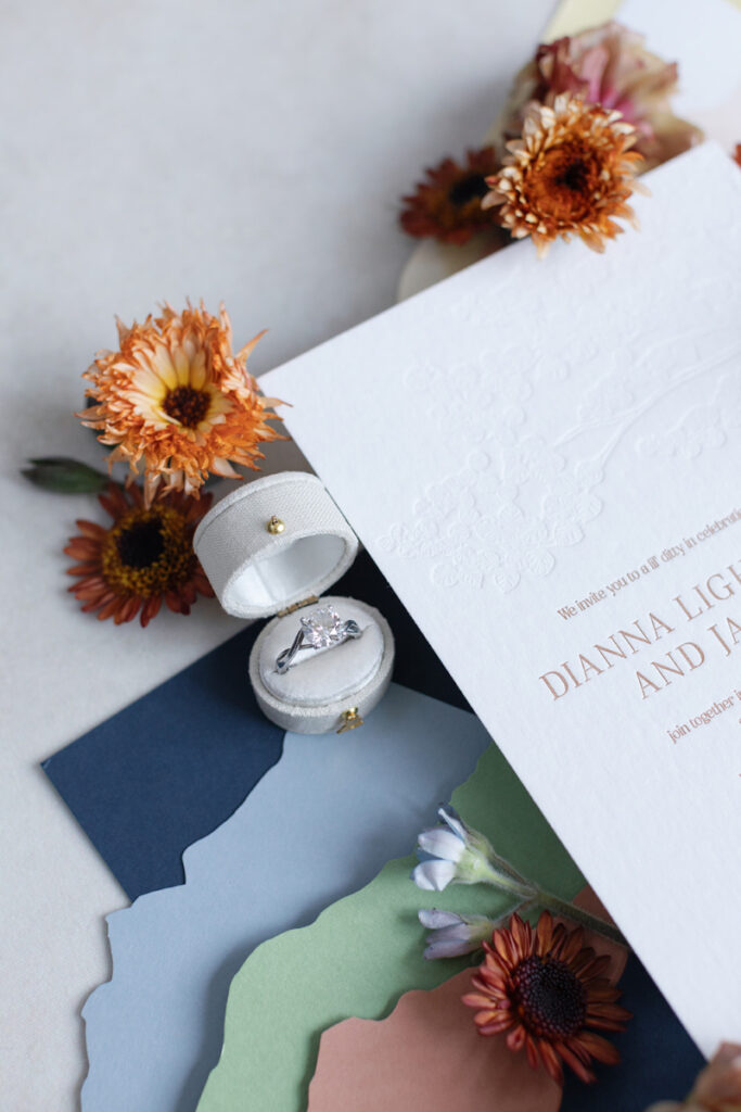 Flatlay with wedding invitation, engagement ring in velvet box, dried florals, and paper details.