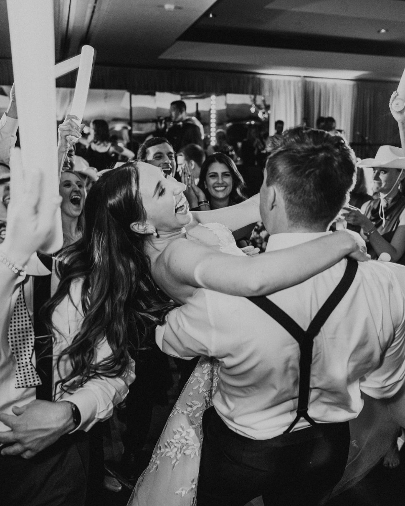 Bride laughing in groom’s arms during a lively dance floor moment surrounded by cheering guests.