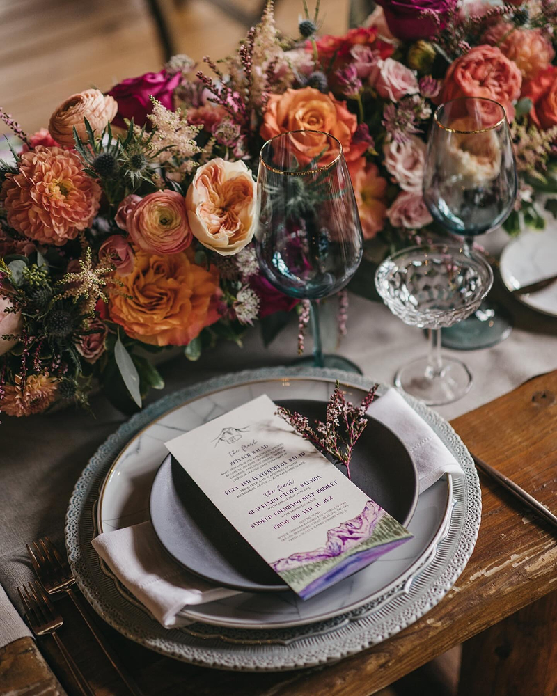 Romantic fall wedding place setting with layered menus, crystal glassware, and lush orange-toned floral arrangement.