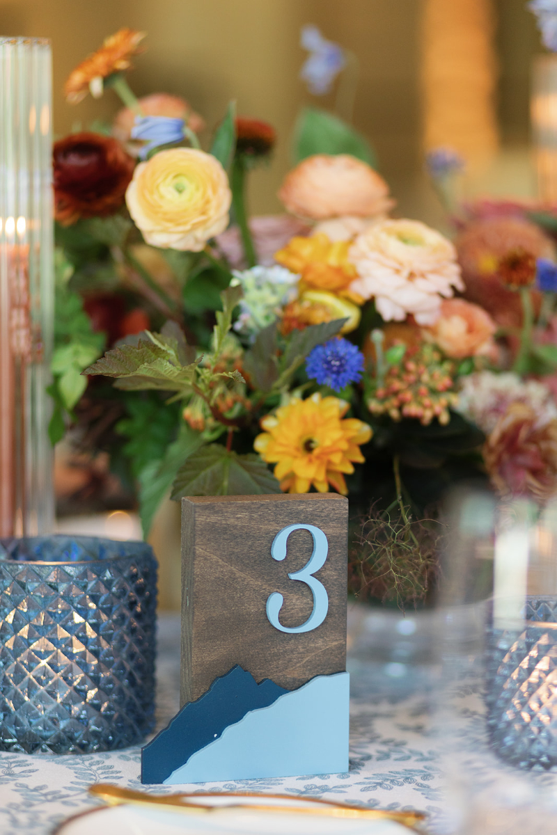 Close-up of a wooden mountain-inspired table number with floral centerpiece and blue glass candle holders.
