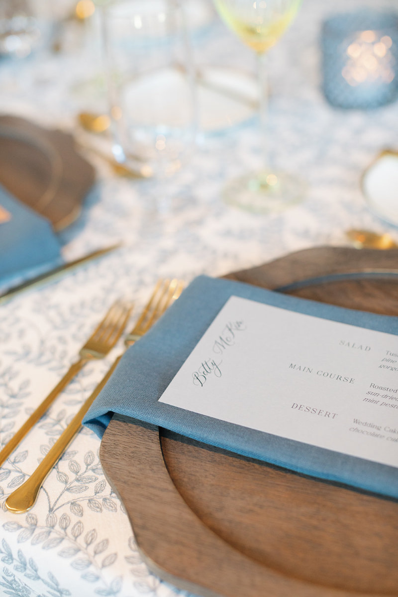 Close-up of a refined place setting with gold flatware, wood charger, blue napkin, and custom menu card.