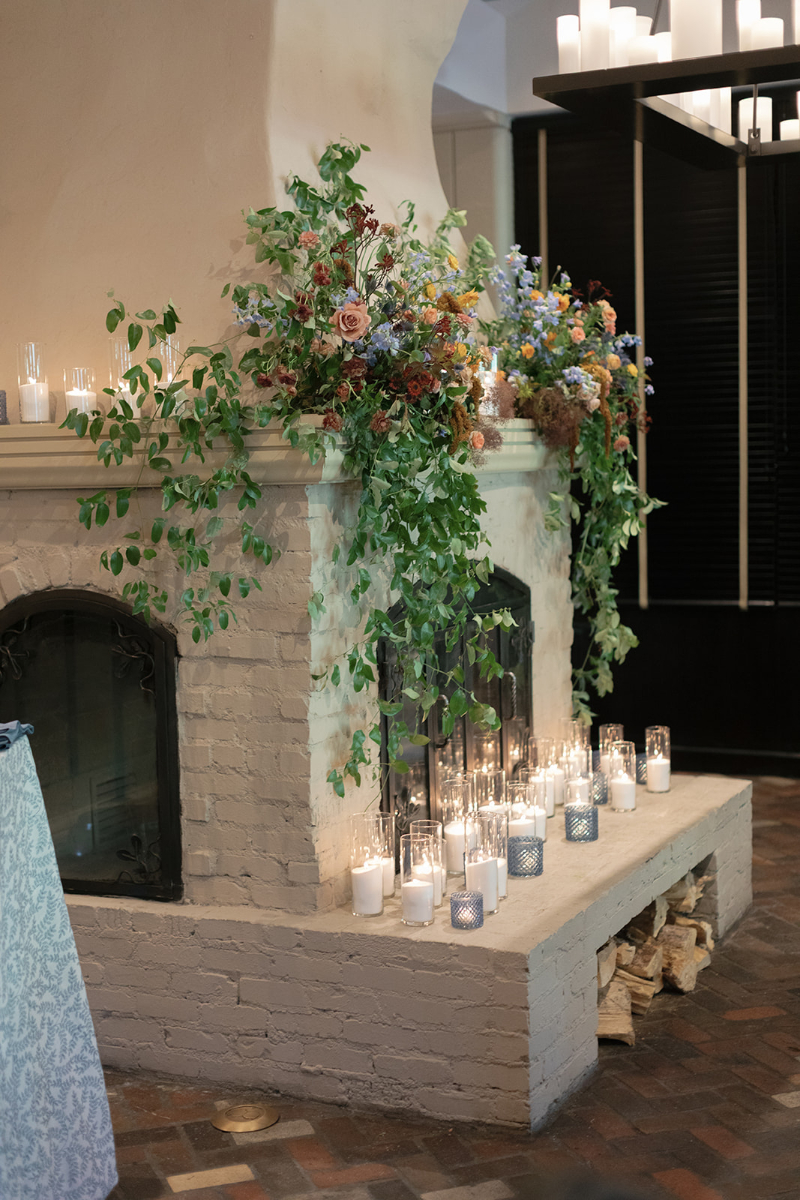 Romantic fireplace floral installation styled by wedding planner with candles, greenery, and soft garden blooms.