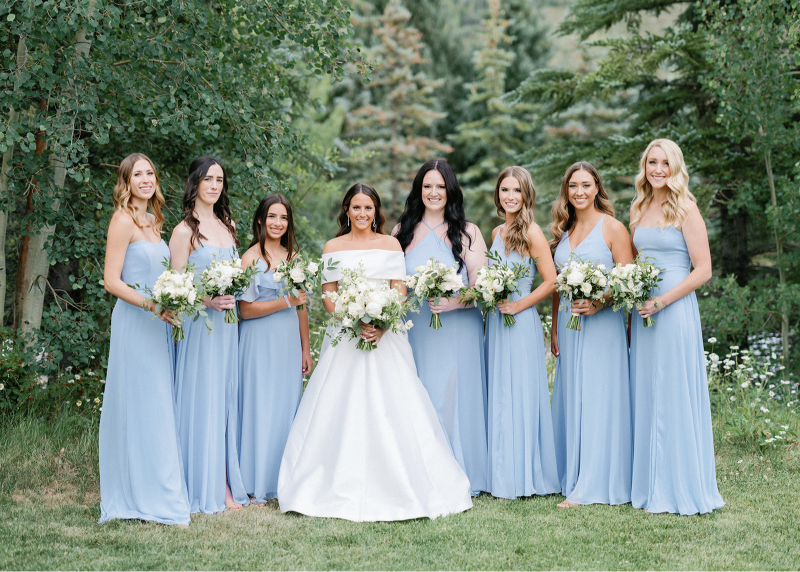 Bride with bridesmaids in light blue dresses holding white bouquets, showcasing cohesive wedding design ideas outdoors.