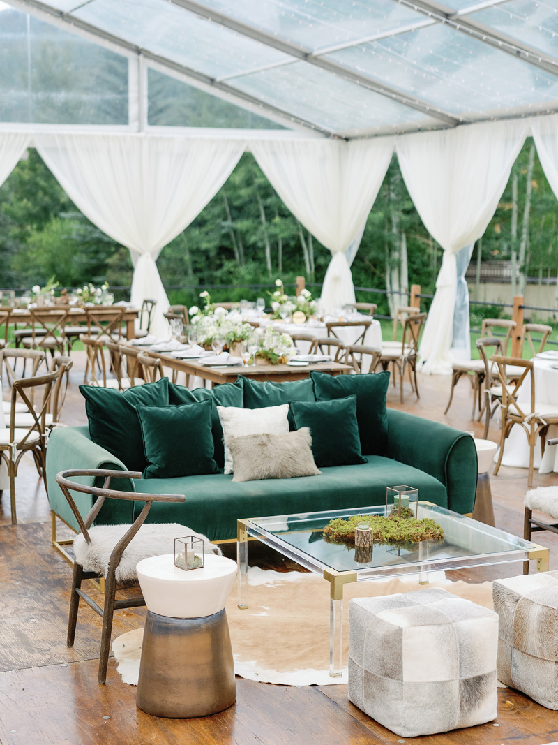 Chic wedding lounge area with emerald green sofa, cowhide rug, modern accents, and forest backdrop.