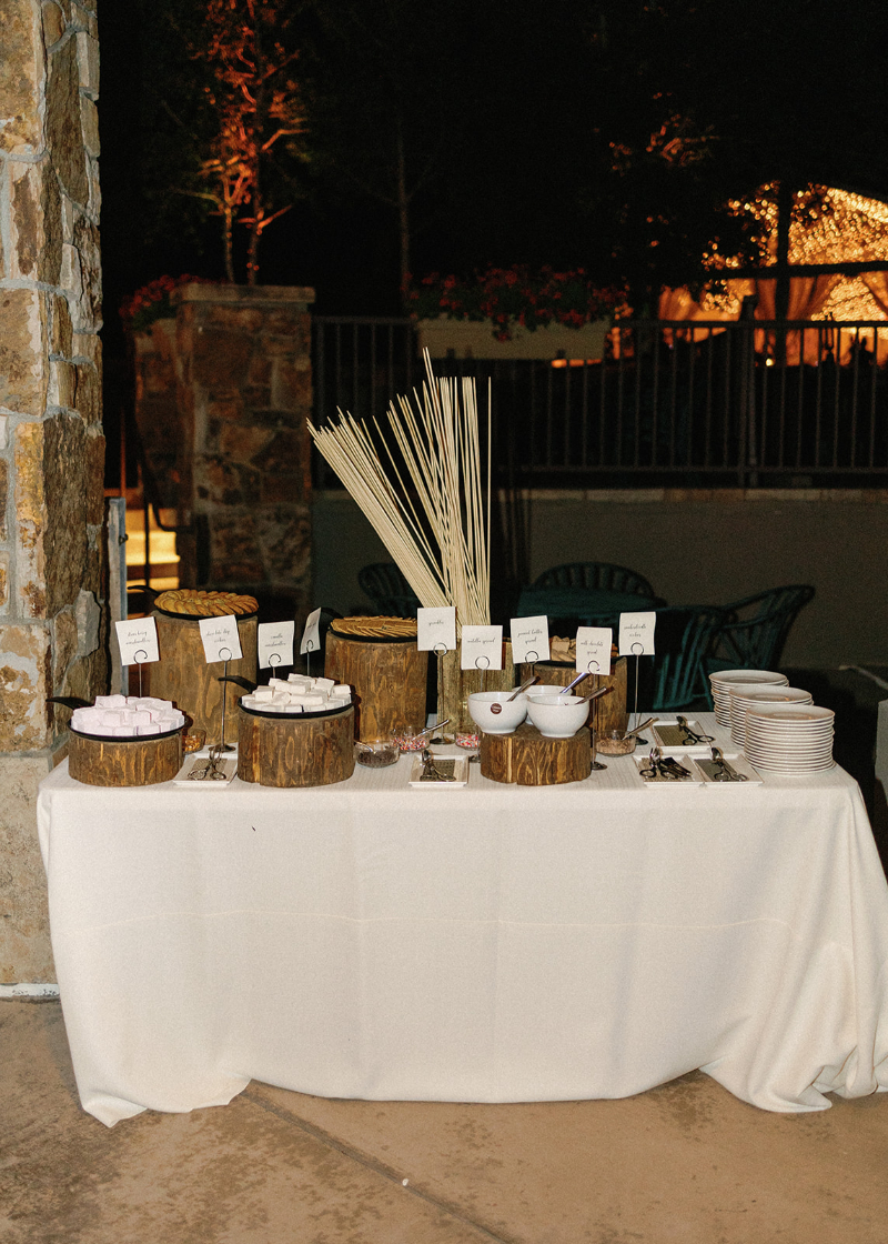 Outdoor s’mores bar with marshmallows, spreads, cookies, skewers, and plates on rustic display.