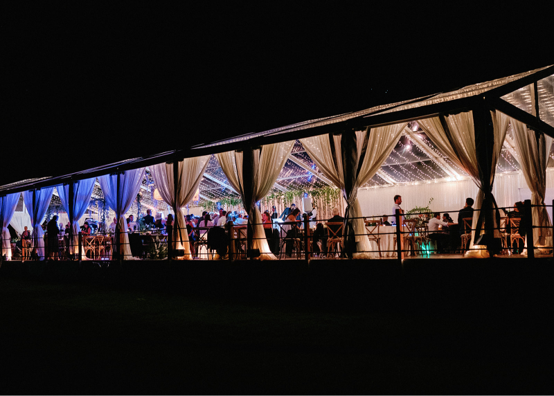 Nighttime wedding reception in clear-top tent glowing with lights, draped in elegant fabric, Colorado wedding design.