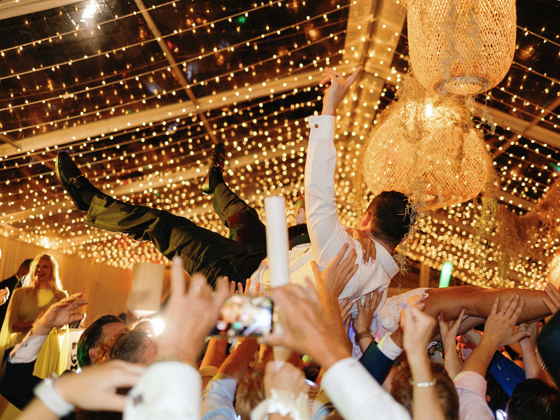 Wedding guest crowd surfs under canopy of twinkle lights and rattan pendants at mountain wedding.