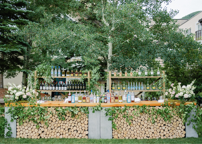 Outdoor wood bar with stacked logs and greenery, showcasing rustic yet refined wedding design ideas.
