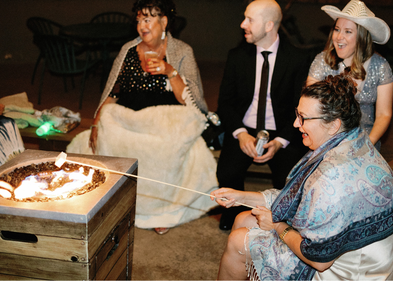 Guests gathered around a firepit at night, roasting marshmallows and enjoying drinks and conversation.