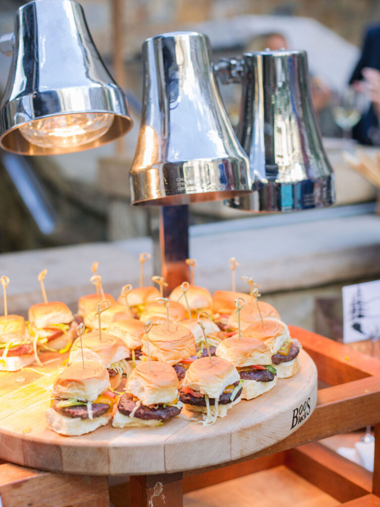 Mini sliders displayed on a wooden board under warming lights at a wedding late-night food station.