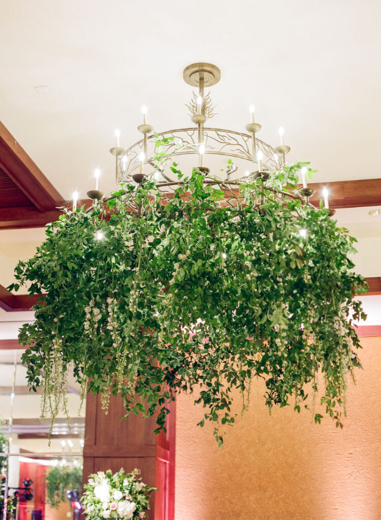 Chandelier wrapped in cascading greenery and soft lighting, suspended in an indoor wedding reception space.