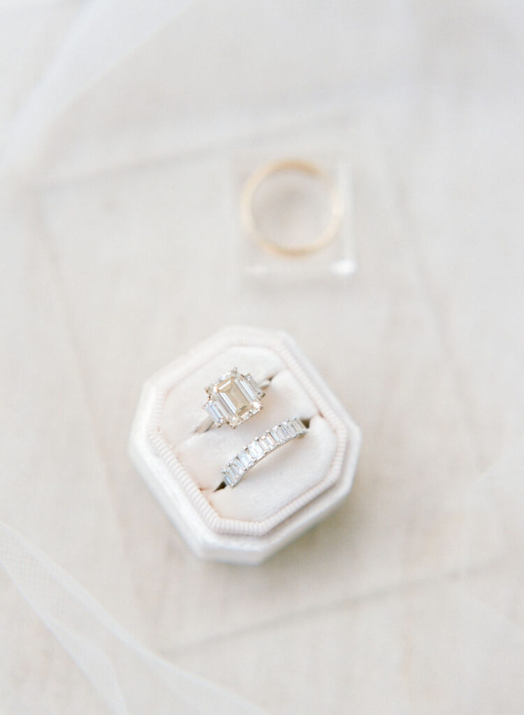 Elegant emerald cut engagement ring and wedding band styled on soft neutral backdrop.