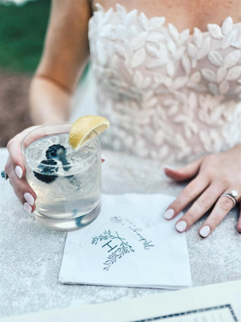 Bride holding signature cocktail with lemon garnish beside custom wedding napkin.