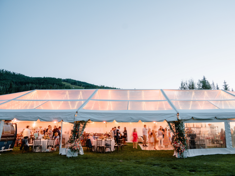 Clear top wedding tent glowing at dusk with guests mingling on mountain lawn.