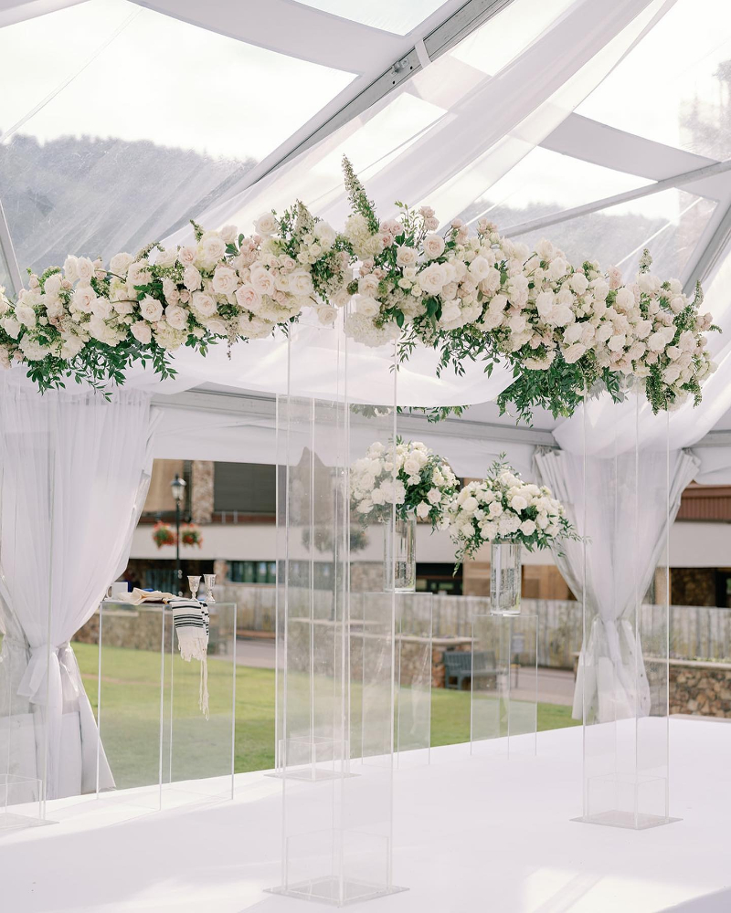 Elegant ceremony setup with clear acrylic chuppah and white floral arrangements under soft draped canopy