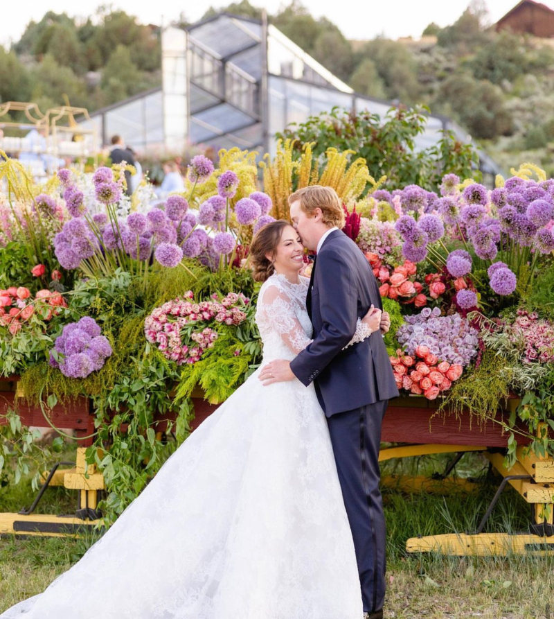 Bride and groom embracing in front of a vibrant floral installation with a glass reception tent in the background.