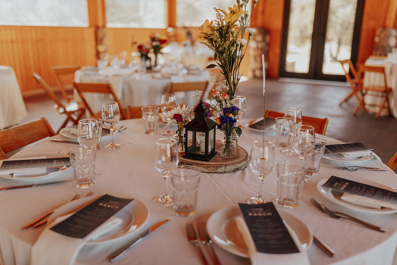 Rustic reception table setting with lantern centerpiece, wildflowers, and neatly arranged place settings inside a warm wood pavilion.