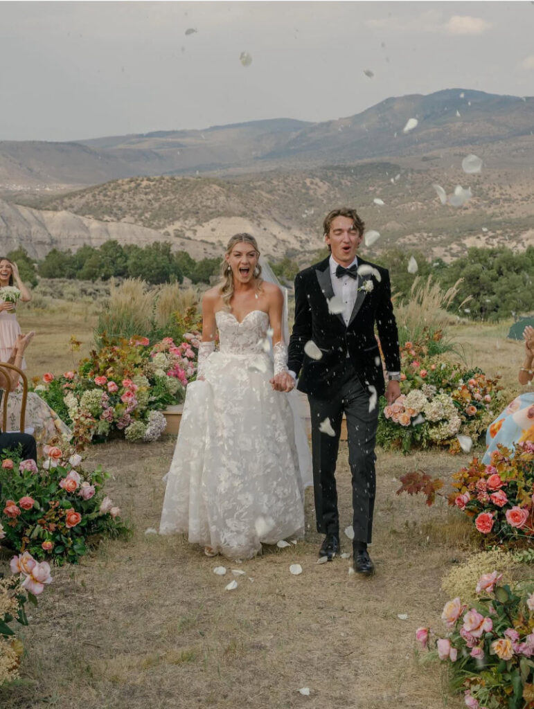 Bride and groom walking down the aisle during a Colorado ranch wedding with mountain views and colorful floral arrangements.