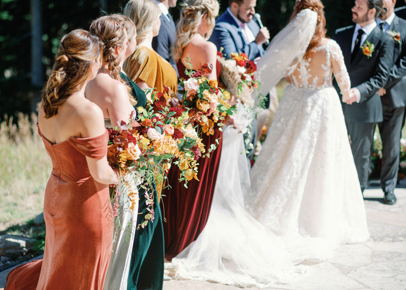Bride and groom exchange vows during an outdoor ceremony surrounded by bridesmaids carrying colorful bouquets.