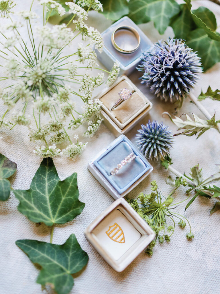 Diamond engagement ring and wedding band in velvet boxes surrounded by delicate white and blue flowers.