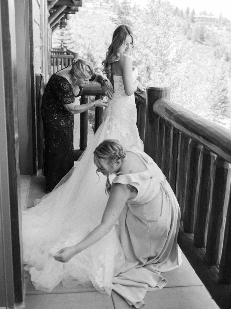 Bride getting ready in her wedding gown with loved ones helping on a rustic balcony.