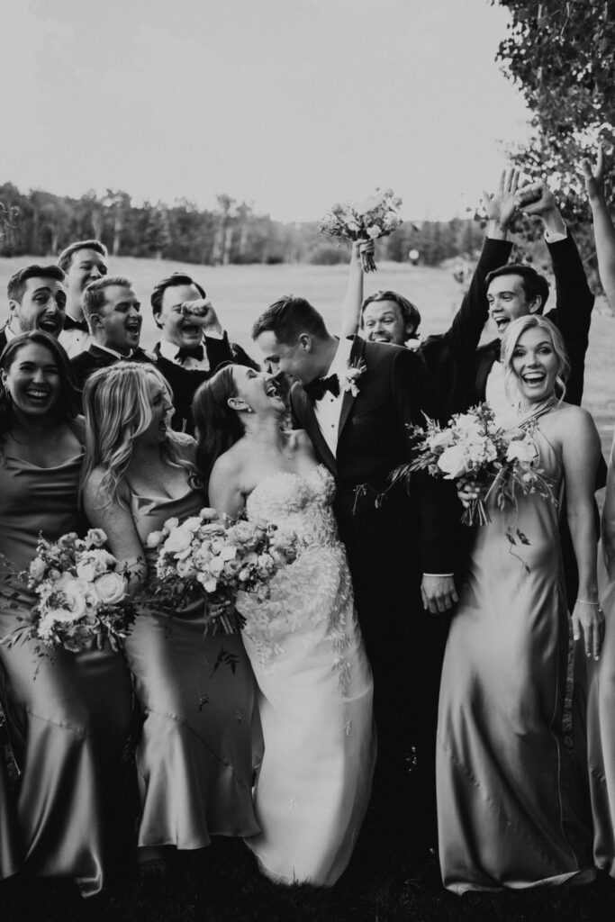 Bride and groom kiss while their wedding party cheers around them during an outdoor celebration.